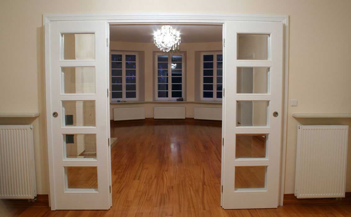 Otwarte przeszklone drzwi wewnętrzne na wymiar w kolorze białym, za nimi duży, nieumeblowany jeszcze salon o półokrągłym kształcie właściwie otoczony drewnianymi oknami (okna również w kolorze białym).