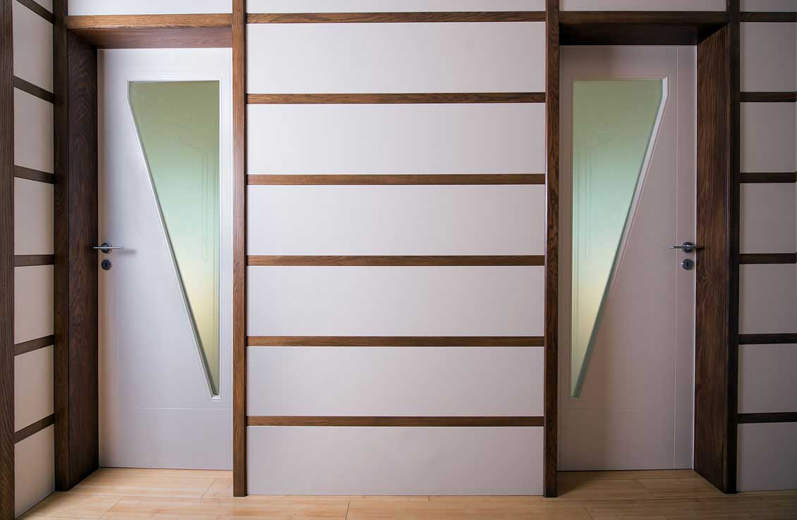 Drewniane drzwi wewnętrzne z przeszkleniami na wymiar, dwie sztuki na jednej ścianie. Biała ściana między drzwiami ozdobiona jest drewnianymi, poziomymi listwami. Przeszklenia w drzwiach mają kształty trójkątów.
