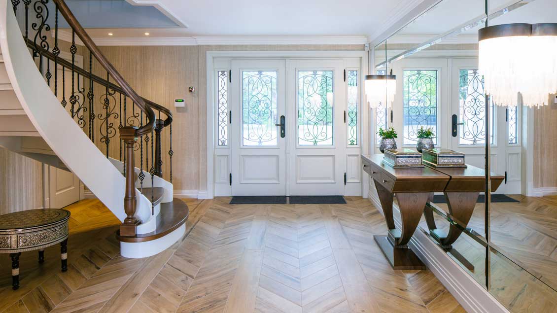 Drzwi zewnętrzne drewniane, częściowo przeszklone - widok z ekskluzywnie urządzonego korytarza. Po bokach widoczne eleganckie drewniane schody i luksusowe drewniane meble na wymiar.