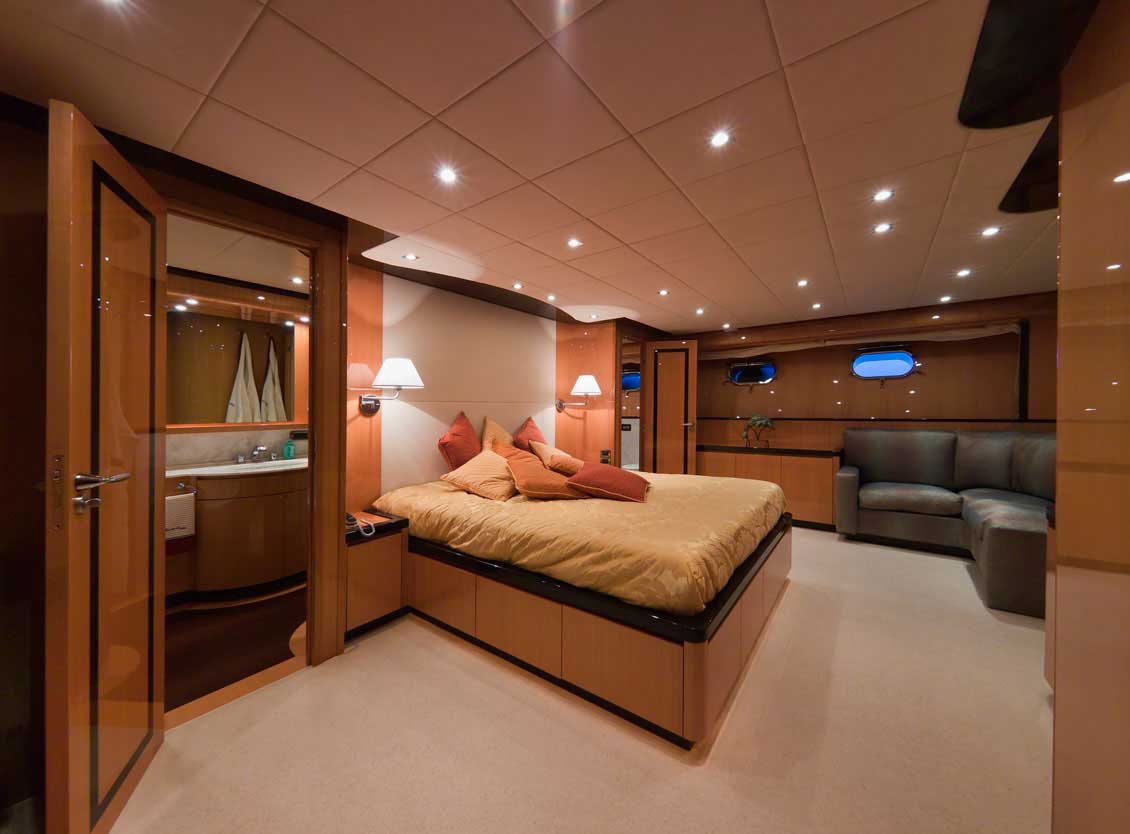 Ekskluzywne drewniane meble na wymiar w prestiżowej kabinie jachtu / statku. Wśród eleganckich mebli i innego wyposażenia na pierwszym planie widoczne są szeroko otwarte dość grube drewniane drzwi i luksusowe łóżko wyprodukowane specjalnie na indywidualne zamówienie na potrzeby tego konkretnego wnętrza.