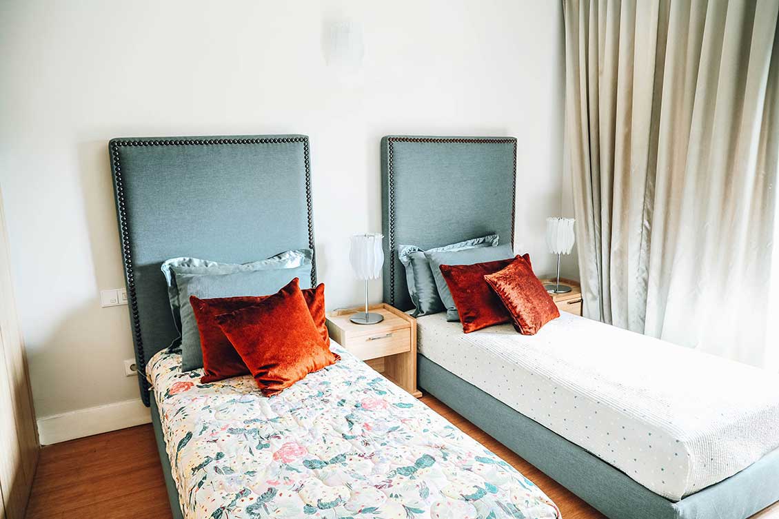 Dwa łóżka pojedyncze z tapicerowanymi zagłowiami w dwuosobowym pokoju domu studenckiego. Obok łóżek małe drewniane stoliki / szafki nocne