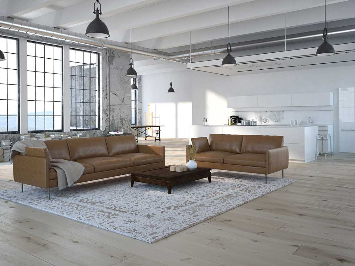 Nowoczesne meble do apartamentu w stylu loft - duże, luksusowe sofy / kanapy i stojący między nimi stolik w kolorze brązowym. W tle zabudowany biały aneks kuchenny.
