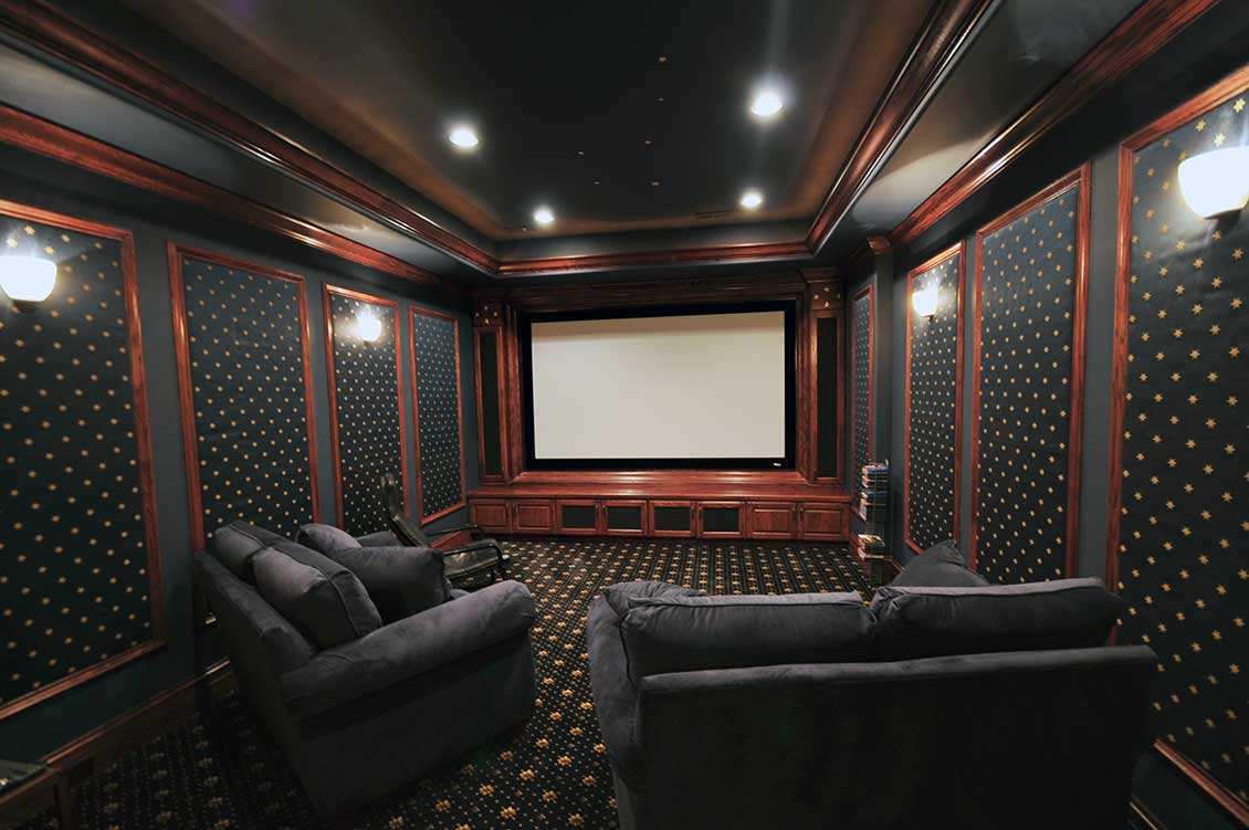 Prywatna, luksusowa sala kinowa, a w niej dwie czarne, luksusowe sofy na wymiar, bardziej z przodu również jeden zdecydowanie mniej luksusowy fotel. Zdjęcie prezentuje widok na ekran z tylnej części sali - zza sof.