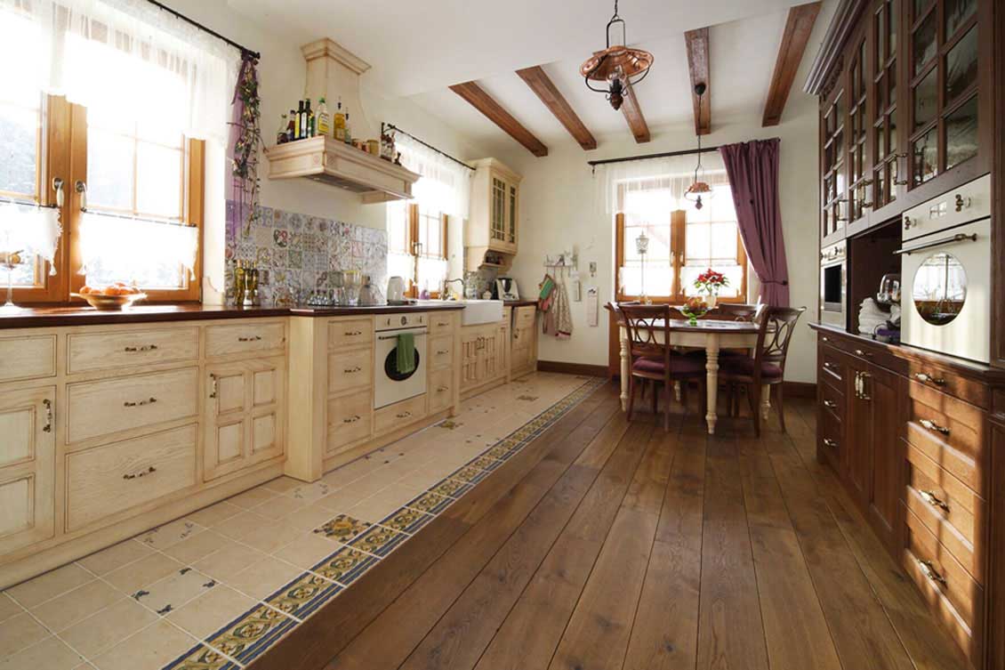 Drewniane meble kuchenne na wymiar. Meble białe po lewej stronie i brązowe po prawej, podłoga większości drewniana, ale częściowo pokryta też kaflami. Kuchnia jest podłużna, w pewnej odległości widać stół i okno.